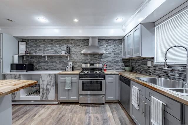 kitchen with appliances with stainless steel finishes, butcher block countertops, sink, gray cabinetry, and wall chimney range hood