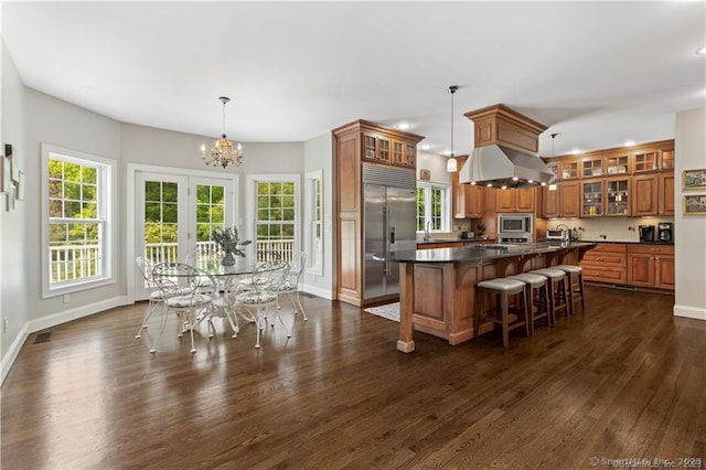 kitchen with dark wood-type flooring, ventilation hood, a center island with sink, built in appliances, and decorative light fixtures