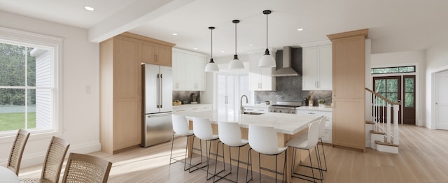 kitchen with high end refrigerator, a center island with sink, wall chimney range hood, decorative backsplash, and decorative light fixtures