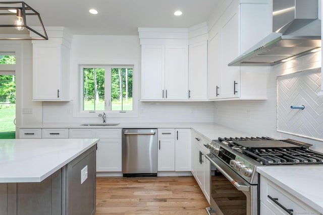 kitchen with wall chimney exhaust hood, sink, pendant lighting, stainless steel appliances, and white cabinets