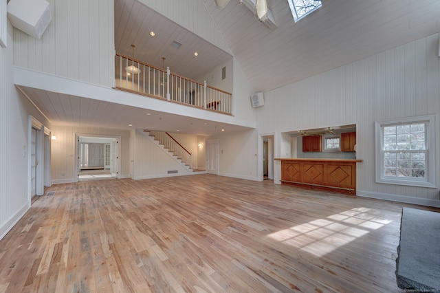 unfurnished living room with high vaulted ceiling, stairway, a skylight, light wood-type flooring, and baseboards