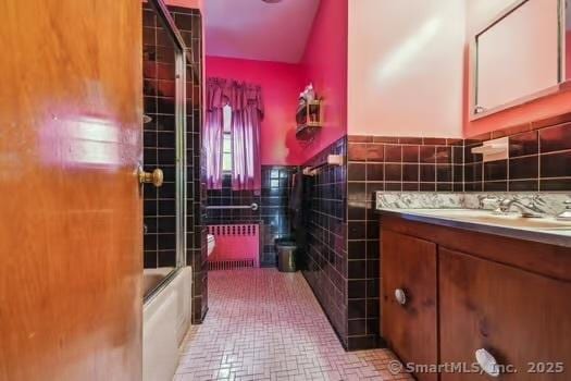 bathroom with radiator, tile patterned flooring, bath / shower combo with glass door, vanity, and tile walls