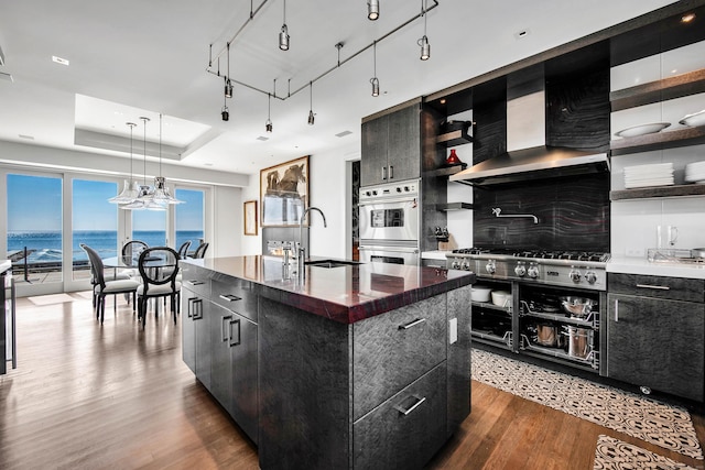 kitchen featuring sink, wall chimney exhaust hood, a water view, an island with sink, and dark wood-type flooring