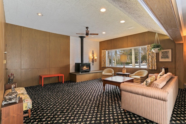 carpeted living room featuring lofted ceiling, a textured ceiling, a wood stove, wooden walls, and ceiling fan