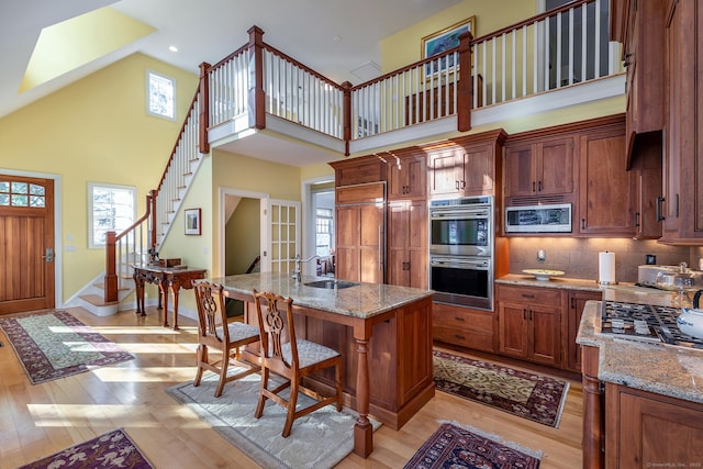 kitchen featuring high vaulted ceiling, an island with sink, built in appliances, light stone counters, and light hardwood / wood-style flooring