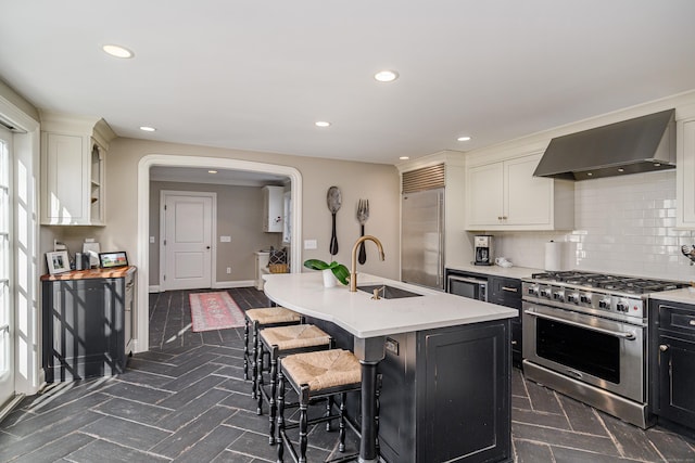 kitchen with sink, a kitchen island with sink, backsplash, premium appliances, and wall chimney exhaust hood