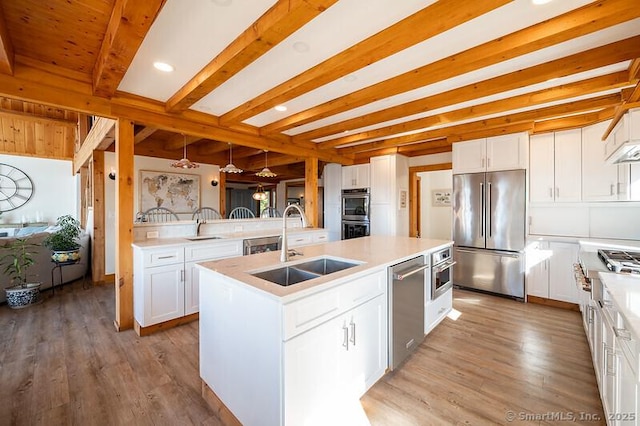 kitchen featuring sink, appliances with stainless steel finishes, white cabinets, a center island with sink, and beamed ceiling