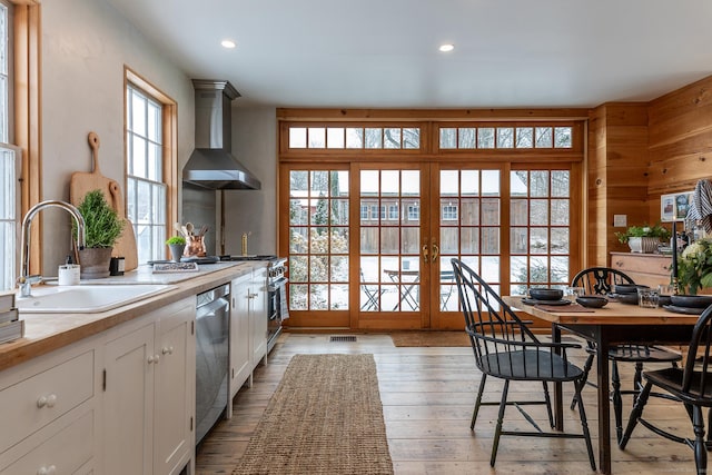 kitchen with white cabinetry, light wood-type flooring, wall chimney range hood, stainless steel dishwasher, and sink