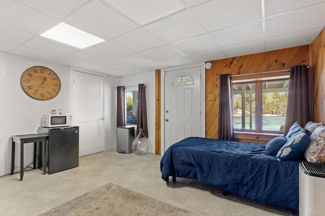 bedroom featuring multiple windows, wooden walls, a drop ceiling, and fridge