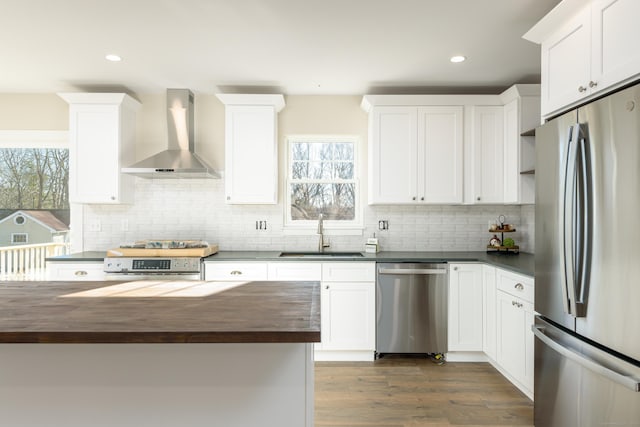 kitchen with appliances with stainless steel finishes, butcher block counters, sink, white cabinets, and wall chimney range hood