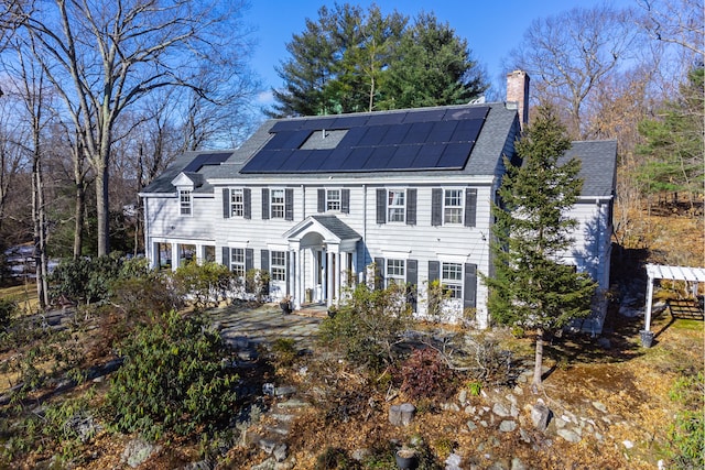 colonial inspired home with a shingled roof, a chimney, and solar panels