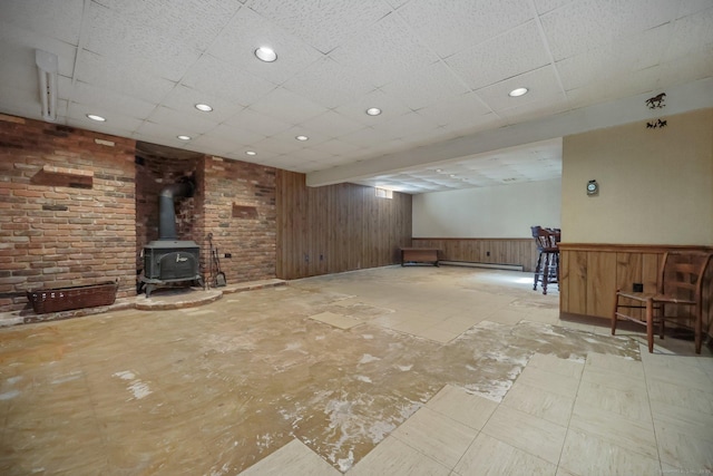 basement with a paneled ceiling, baseboard heating, wooden walls, brick wall, and a wood stove