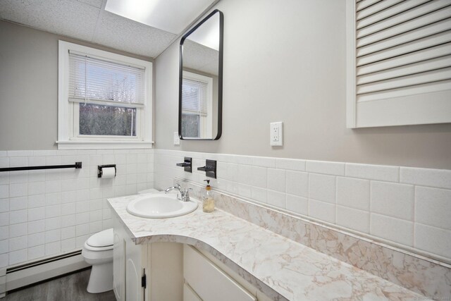 bathroom with hardwood / wood-style flooring, baseboard heating, tile walls, vanity, and toilet