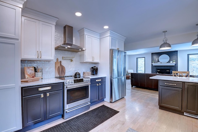 kitchen with stainless steel appliances, white cabinets, a brick fireplace, decorative light fixtures, and wall chimney exhaust hood