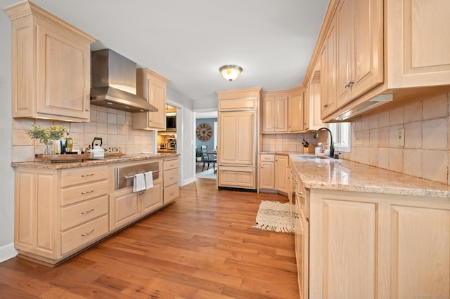 kitchen with wall chimney exhaust hood, light brown cabinetry, sink, light stone counters, and light wood-type flooring