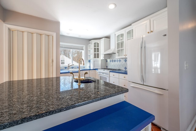 kitchen with white appliances, white cabinetry, wall chimney range hood, dark stone counters, and decorative backsplash