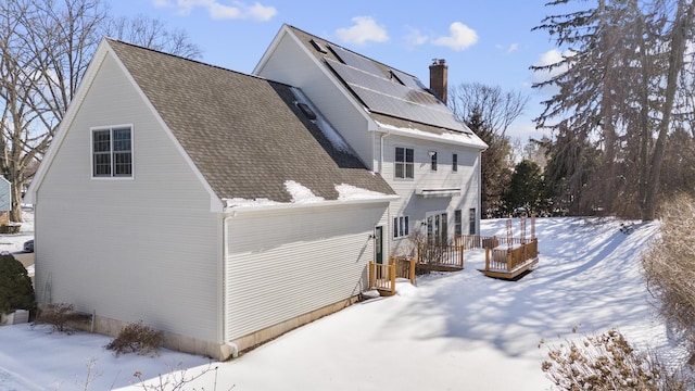 view of snow covered exterior featuring a shingled roof, roof mounted solar panels, a chimney, and a wooden deck