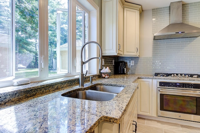 kitchen with appliances with stainless steel finishes, wall chimney exhaust hood, cream cabinetry, tasteful backsplash, and light stone counters