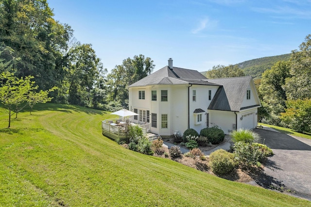 rear view of property with aphalt driveway, roof with shingles, a lawn, a deck, and a garage