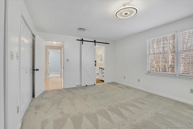 unfurnished bedroom featuring ensuite bathroom, a barn door, light carpet, and a textured ceiling