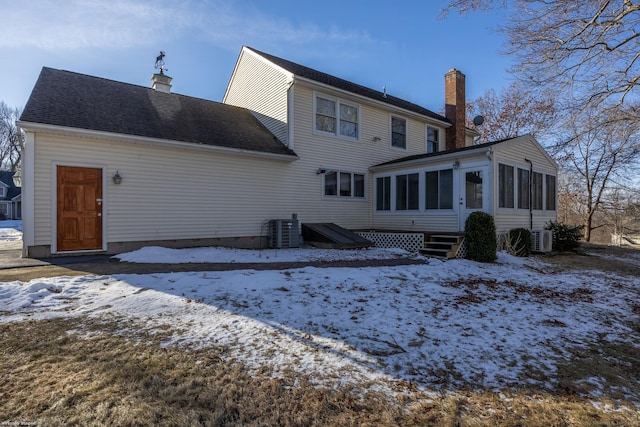 snow covered house featuring entry steps, central AC unit, a chimney, and a sunroom