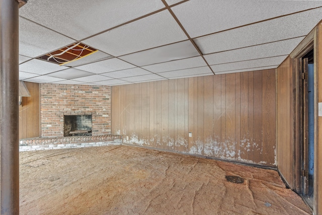 unfurnished living room with a brick fireplace, carpet flooring, a drop ceiling, and wooden walls