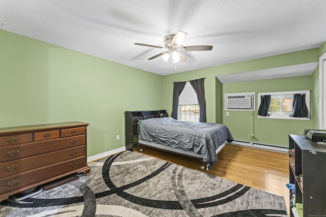 bedroom with ceiling fan, baseboard heating, hardwood / wood-style floors, a wall unit AC, and a textured ceiling