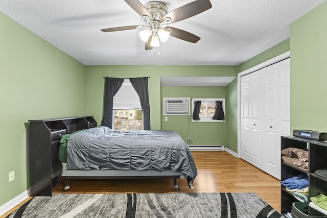 bedroom featuring a wall mounted air conditioner, wood-type flooring, a baseboard radiator, ceiling fan, and a closet