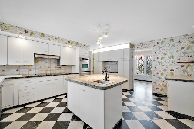 kitchen with dark floors, white cabinets, a sink, an island with sink, and wallpapered walls