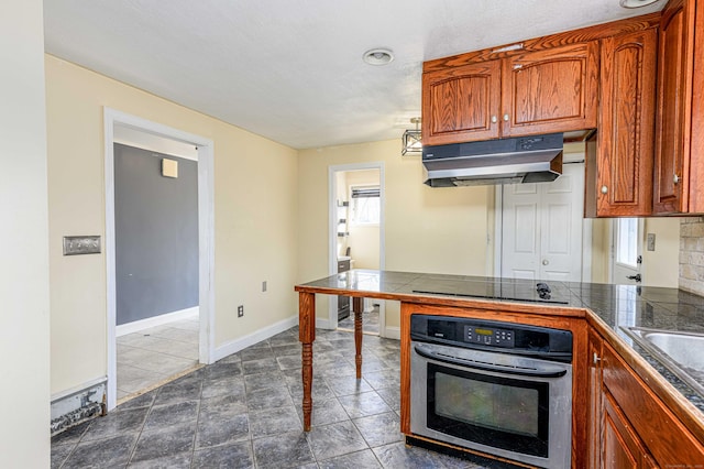 kitchen featuring oven, baseboards, black electric stovetop, under cabinet range hood, and brown cabinetry