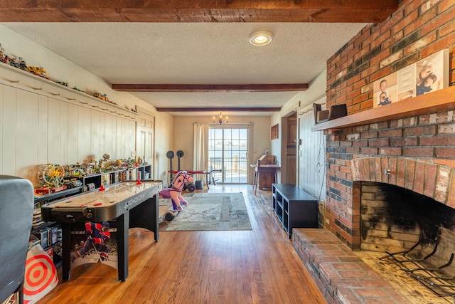 game room with a brick fireplace, a textured ceiling, a notable chandelier, beam ceiling, and hardwood / wood-style floors