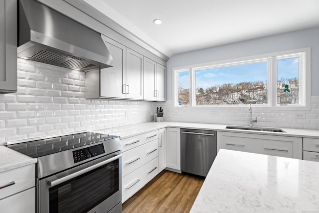 kitchen featuring appliances with stainless steel finishes, wood-type flooring, sink, light stone countertops, and wall chimney range hood