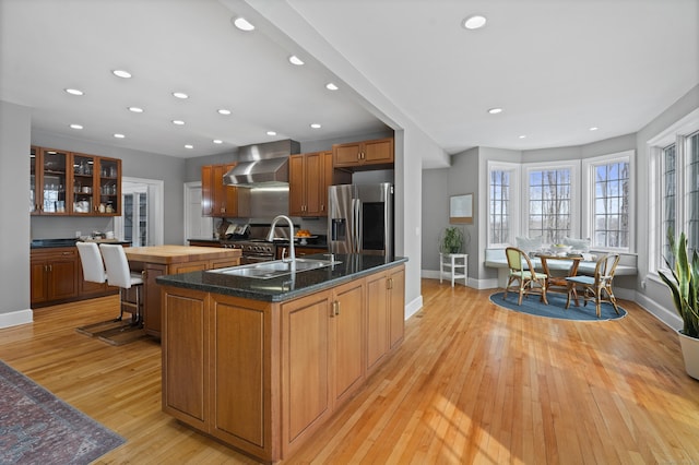 kitchen featuring a sink, stainless steel refrigerator with ice dispenser, wall chimney exhaust hood, light wood finished floors, and an island with sink