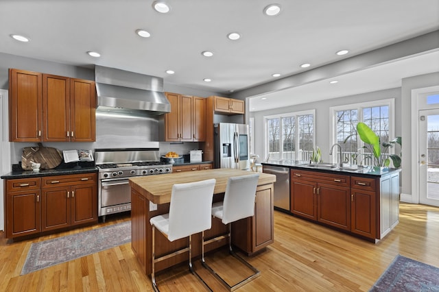 kitchen with a center island with sink, stainless steel appliances, light wood-type flooring, wall chimney range hood, and a sink