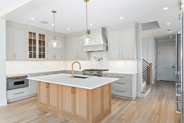 kitchen featuring sink, tasteful backsplash, light hardwood / wood-style floors, custom range hood, and an island with sink