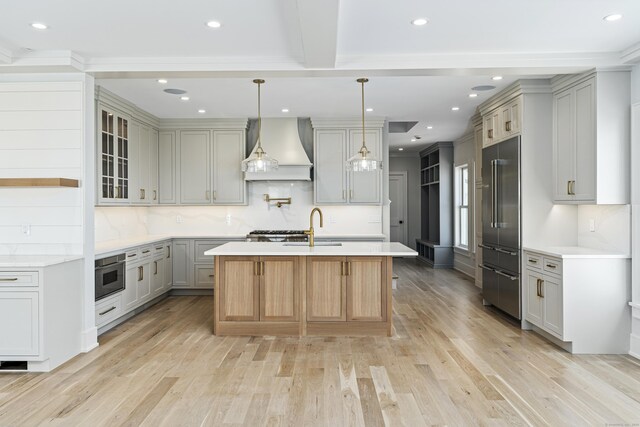 kitchen with a sink, an island with sink, light wood-type flooring, custom exhaust hood, and high quality fridge