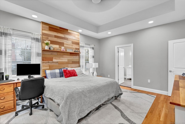 bedroom with ensuite bathroom, wooden walls, a raised ceiling, and light hardwood / wood-style floors