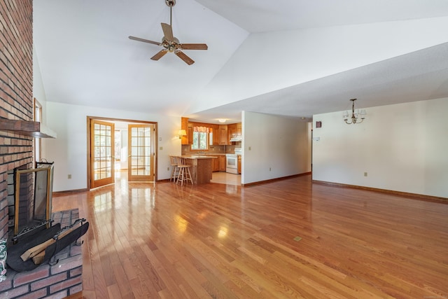 unfurnished living room with a brick fireplace, ceiling fan with notable chandelier, high vaulted ceiling, and light hardwood / wood-style flooring