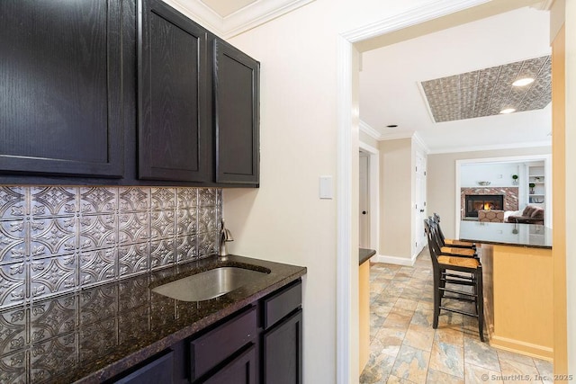 kitchen with sink, a large fireplace, decorative backsplash, dark stone counters, and ornamental molding