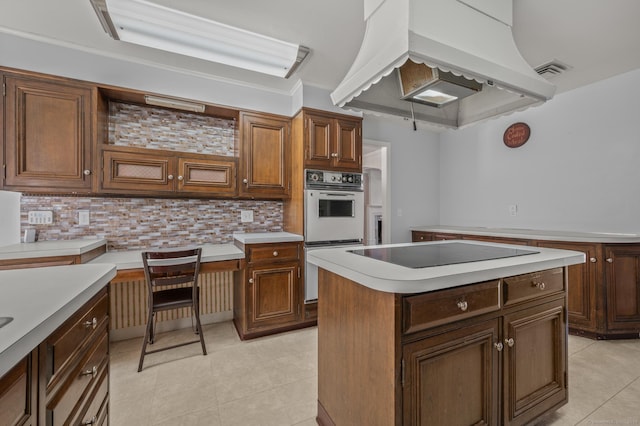 kitchen featuring custom exhaust hood, built in desk, black electric cooktop, double oven, and a kitchen island