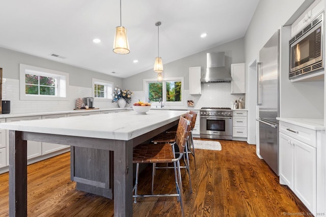 kitchen with white cabinetry, hanging light fixtures, a kitchen breakfast bar, built in appliances, and wall chimney exhaust hood