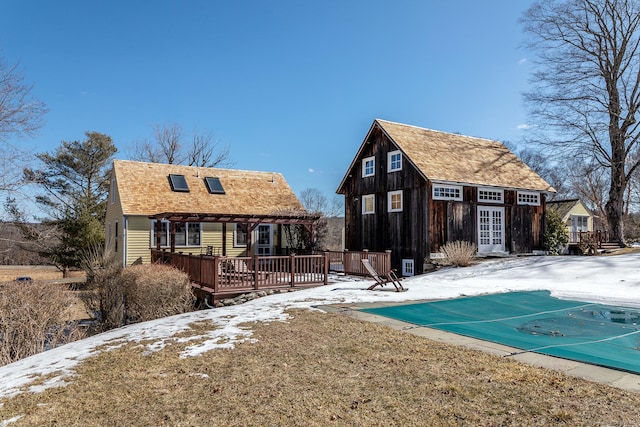 snow covered back of property featuring an outdoor structure, roof with shingles, a wooden deck, a covered pool, and an exterior structure