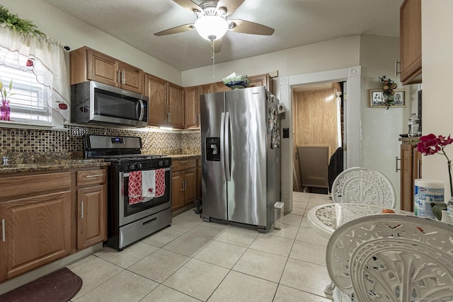 kitchen featuring light tile patterned floors, dark stone countertops, ceiling fan, stainless steel appliances, and backsplash