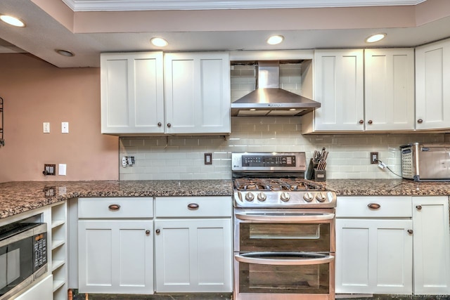 kitchen featuring wall chimney exhaust hood, white cabinetry, dark stone countertops, appliances with stainless steel finishes, and decorative backsplash