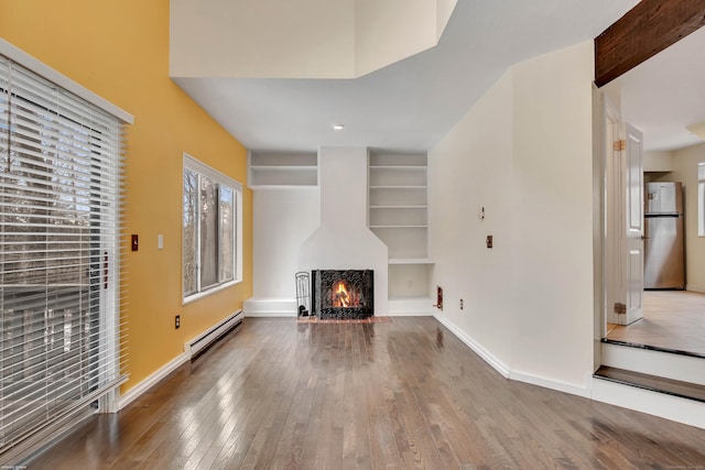 unfurnished living room featuring built in shelves, baseboards, a baseboard radiator, a warm lit fireplace, and hardwood / wood-style floors