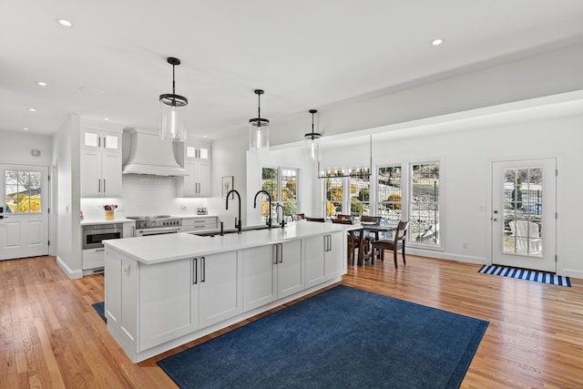 kitchen featuring an island with sink, white cabinetry, decorative light fixtures, sink, and custom exhaust hood