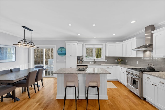 kitchen with wall chimney exhaust hood, white cabinetry, stainless steel stove, a kitchen island, and light stone countertops