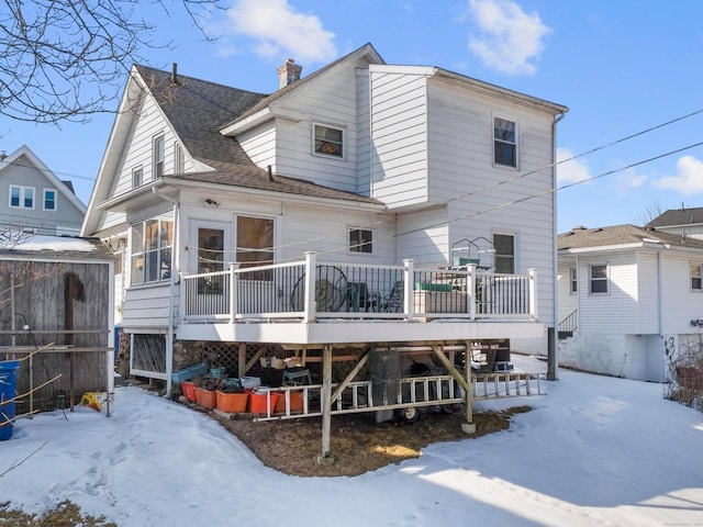 snow covered back of property with a deck, roof with shingles, and a chimney