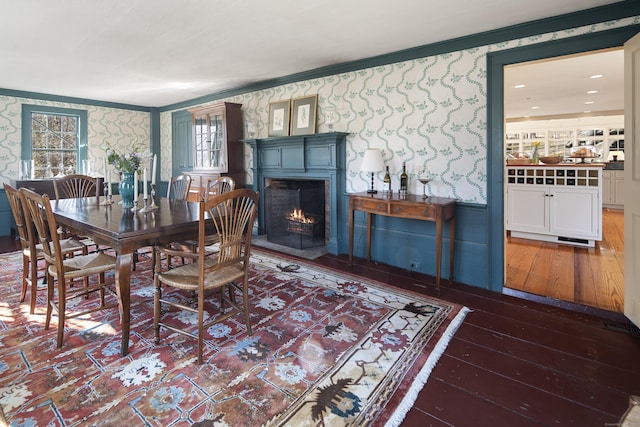 dining room with wallpapered walls, a warm lit fireplace, ornamental molding, and wood-type flooring