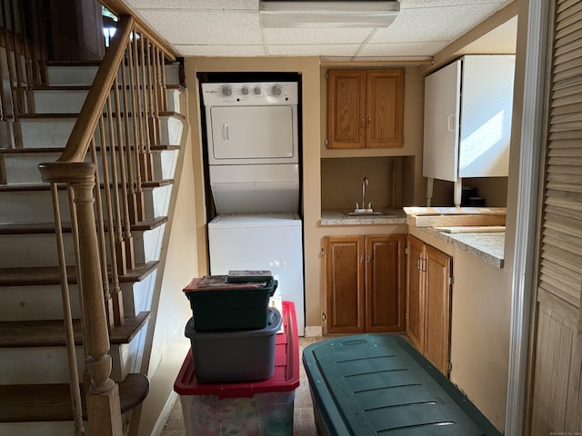 kitchen featuring a paneled ceiling, stacked washer / dryer, a sink, light countertops, and brown cabinetry